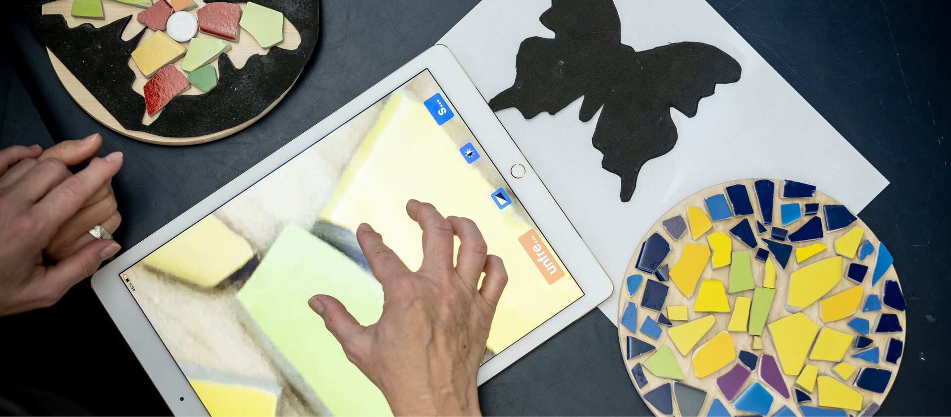 Des mains travaillent avec des pièces de mosaïque colorées, un pochoir noir en forme de papillon et une tablette sur une table.