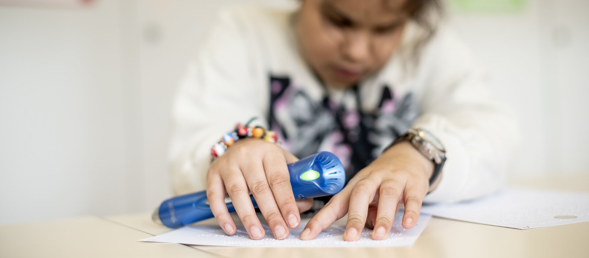 Un enfant parcourt du bout des doigts un texte en braille. Il tient un lecteur bleu dans la main.