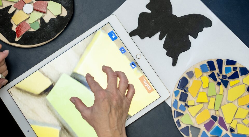 Des mains travaillent avec des pièces de mosaïque colorées, un pochoir noir en forme de papillon et une tablette sur une table.