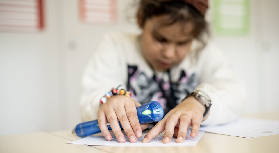 Un enfant parcourt du bout des doigts un texte en braille. Il tient un lecteur bleu dans la main.